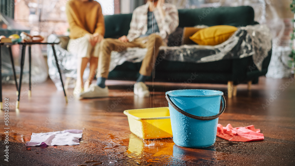 Two homeowners sitting on a couch in the background, in the foreground an active leak dripping into buckets on the living room floor.
