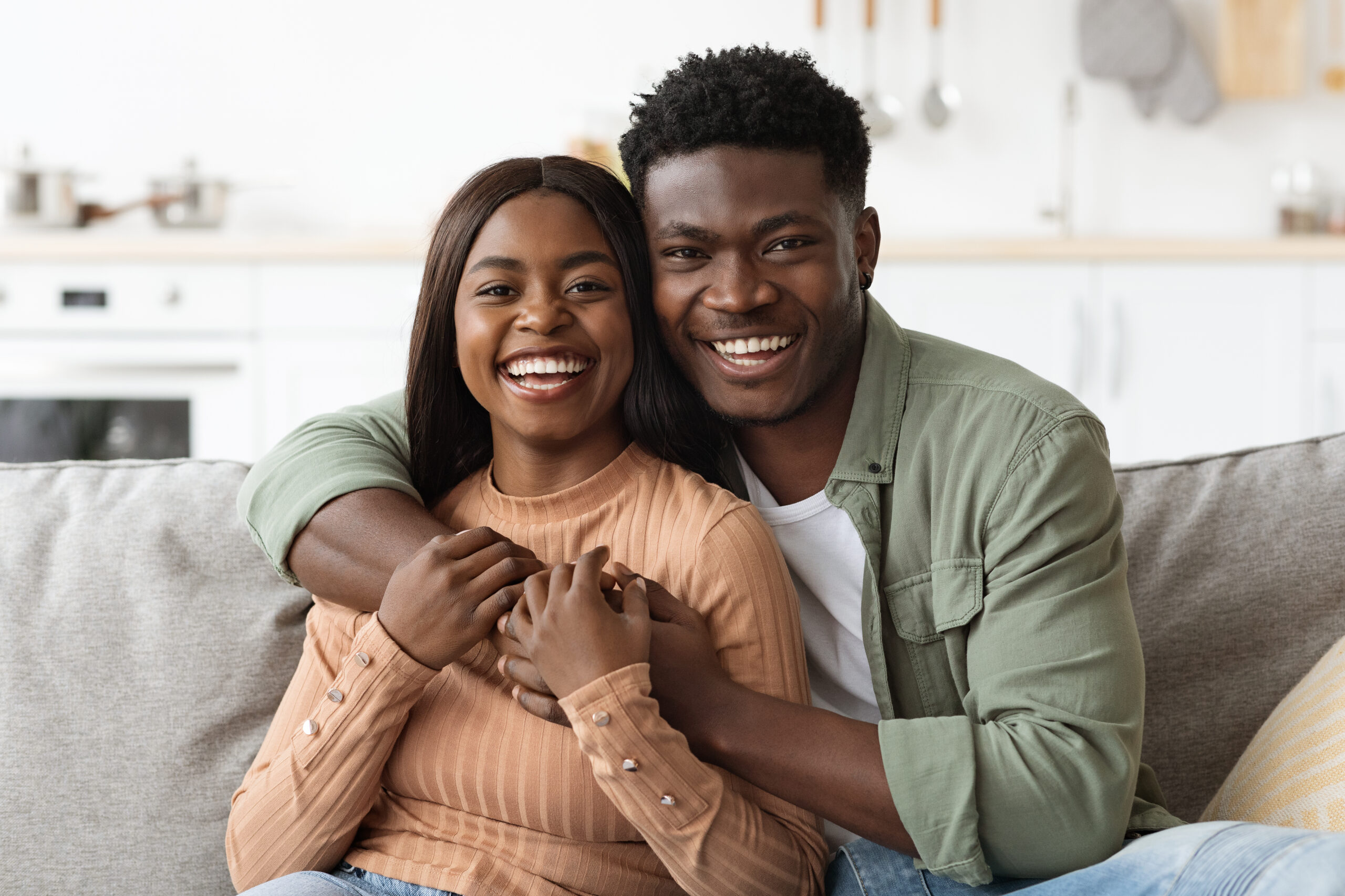 Closeup portrait of happy black couple embracing and smiling at camera, loving man and woman sitting on couch in living room, enjoying weekend together.