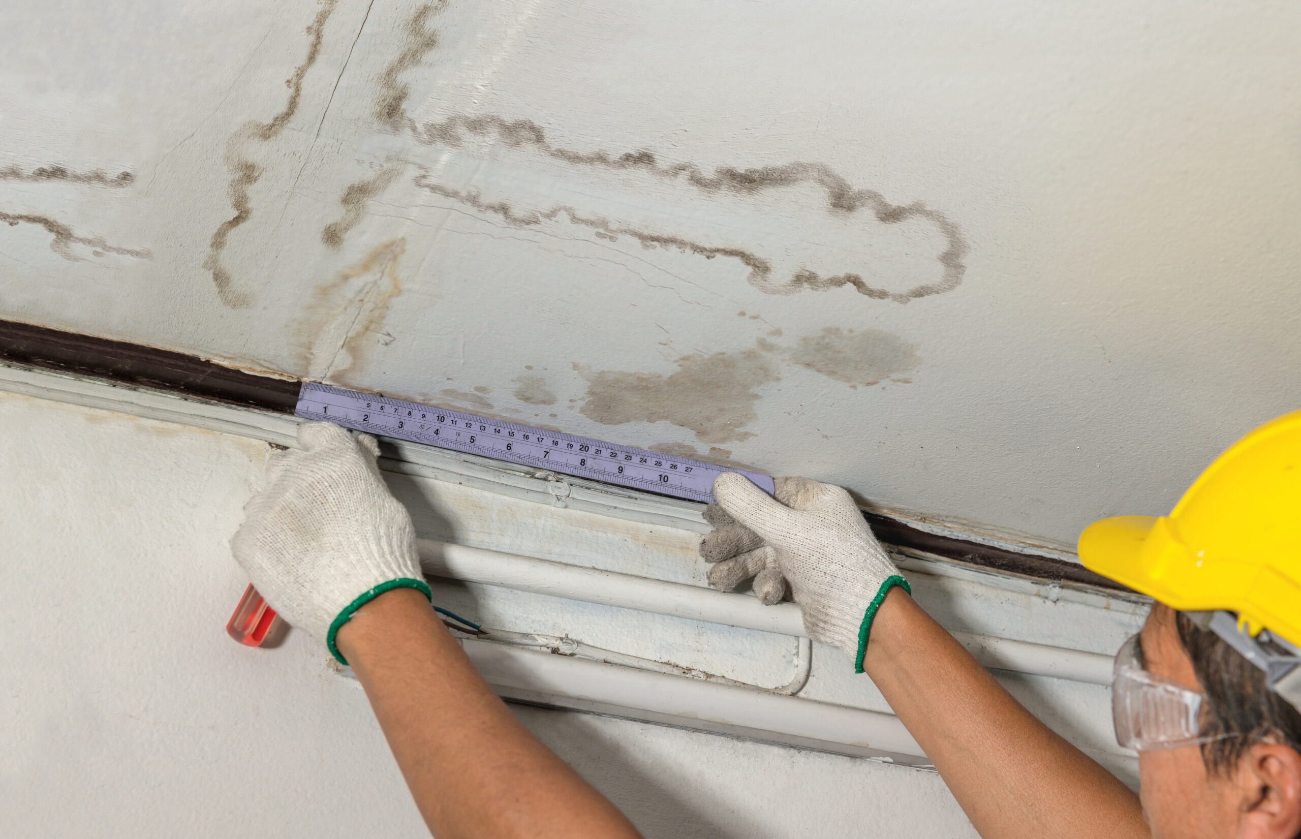 Contractor is measuring a water stain on a ceiling using a ruler. Wearing a yellow safety hat, gloves, and glasses, fully donned in safety gear.