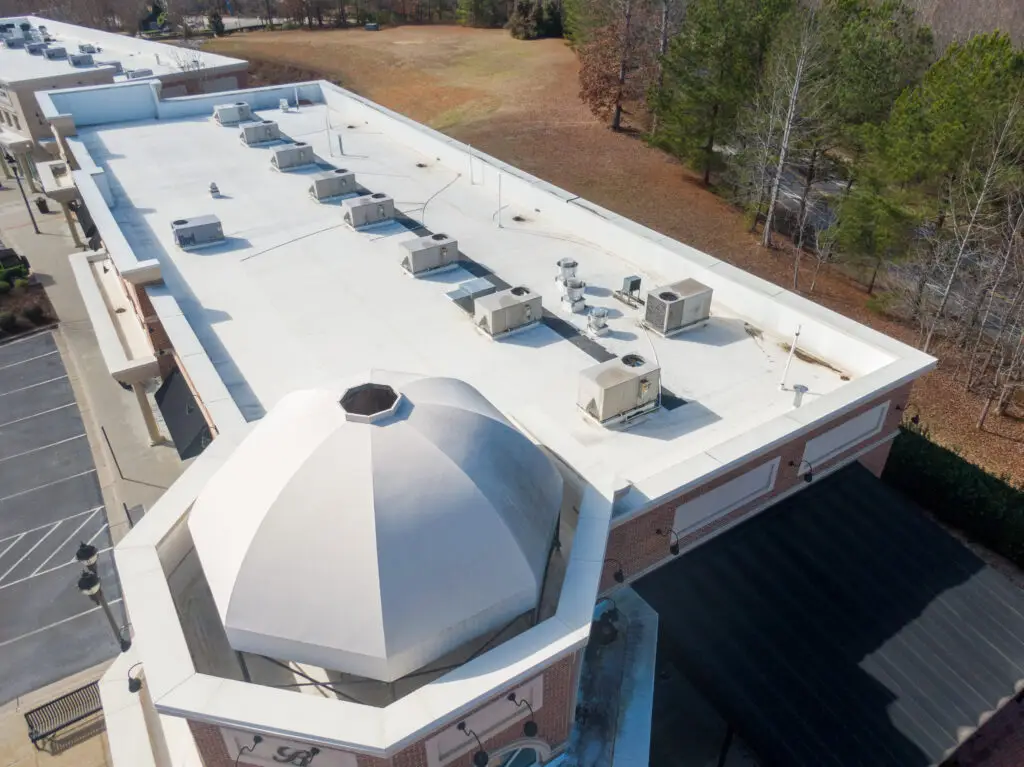An aerial view of a flat commercial roof with white membrane roofing, HVAC units, and a dome-shaped architectural feature, surrounded by trees and parking spaces.