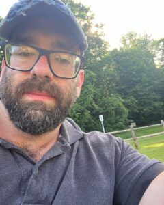 Bearded man in glasses taking a selfie on his property.